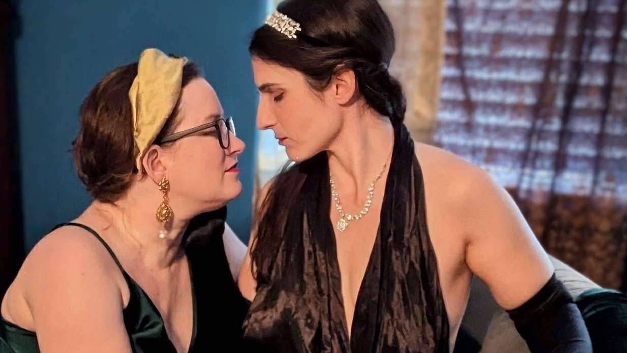 A woman in a 1930s style black dress leans in towards a woman in a green dress and gently caresses her face as both close their eyes.